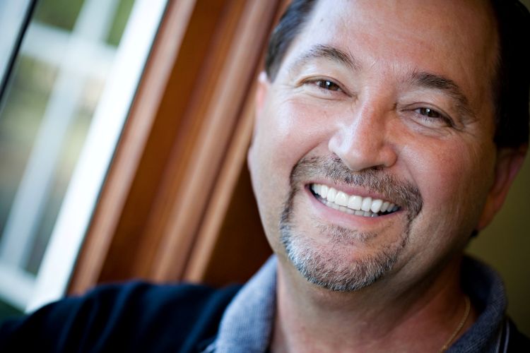 An older man with a tan complexion and dark hair, smiling broadly with his head tilted.