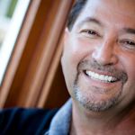 An older man with a tan complexion and dark hair, smiling broadly with his head tilted.