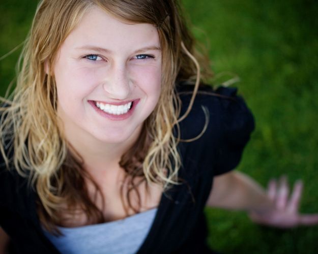 A young woman with blonde hair smiling while standing outdoors in front of greenery.