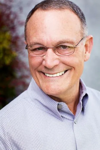 An older man with glasses and a light-colored button-down shirt, smiling.