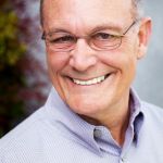An older man with glasses and a light-colored button-down shirt, smiling.