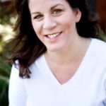 Portrait of a woman with dark wavy hair wearing a white V-neck shirt, smiling warmly.