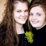 Two young girls with long brown hair posing closely together, both smiling.