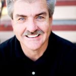 Portrait of an older man with short grey hair, wearing a black shirt and smiling.